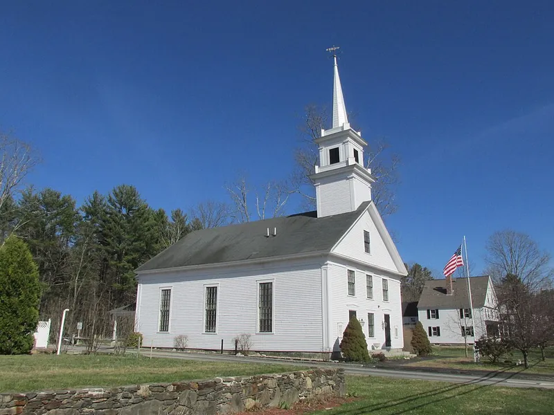 White wooden church with a tall steeple under a bright blue sky; American flag on a nearby flagpole.