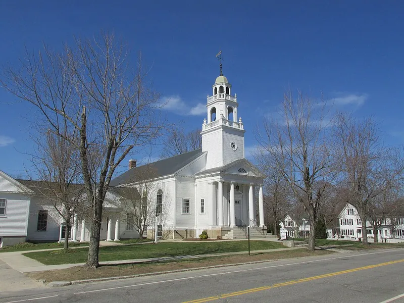 White church with a tall bell tower and columned portico on a street, under a clear blue sky.