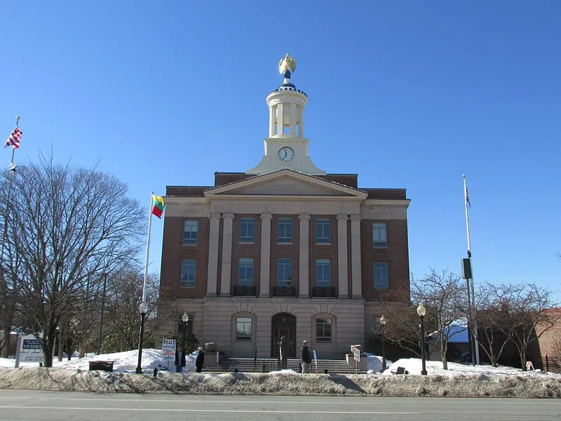 Front view of a brick municipal building with a central clock tower topped by a golden eagle statue, flagpoles on either side, and a clear blue sky behind.