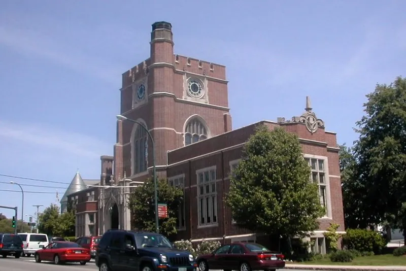 Brick, castle-style building with a tall square tower and arched windows, set on a street with parked cars and trees nearby.