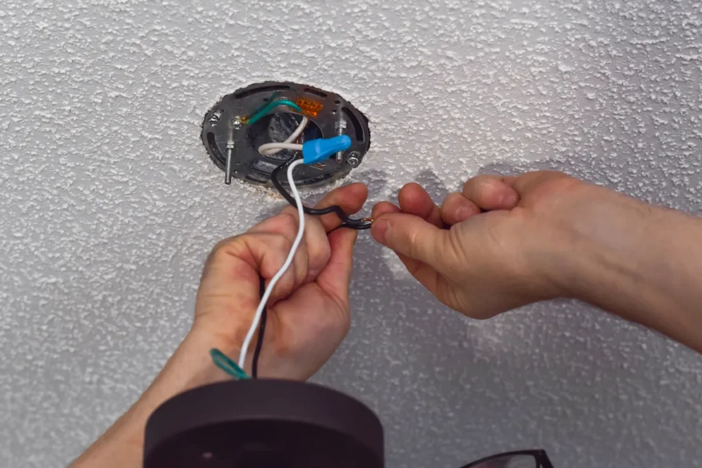 Person wires a ceiling light fixture: black and white conductors joined inside an electrical box with a blue connector nearby.